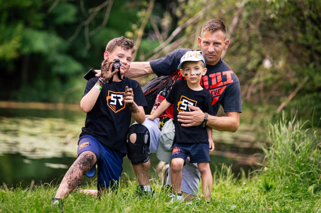 Dwoje dzieci i tata w koszulkach Survival Race Kids pozują do zdjęcia, pokazując ubrudzone błotem ręce po biegu.